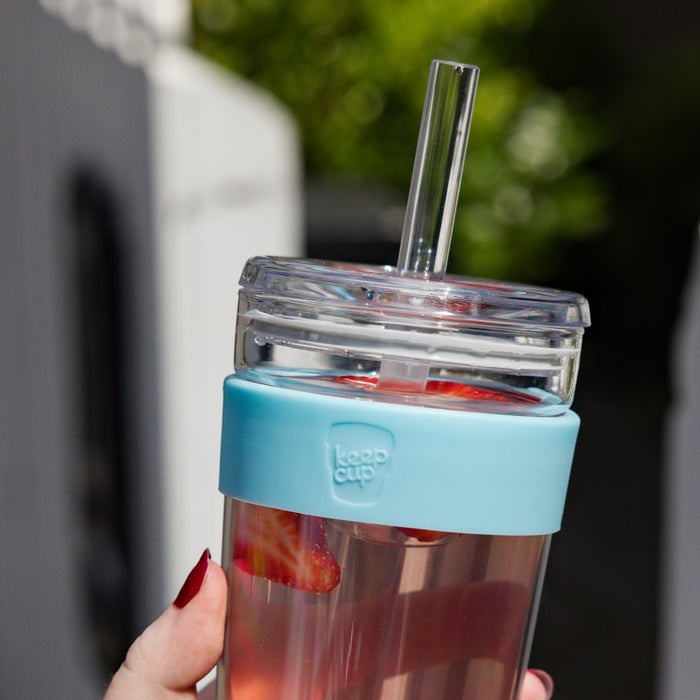 Hand with dark red nail polish holding a KeepCup Cold Cup Longplay Boba M 12oz in Cloud, featuring a light blue silicone band and clear lid with reusable straw, filled with a pink beverage and strawberry pieces, with blurred outdoor greenery in the background.