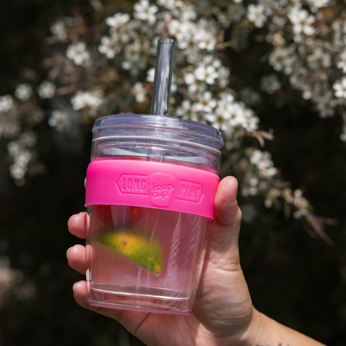 Hand holding a KeepCup Cold Cup Longplay Boba M 12oz in Flamingo, featuring a clear plastic cup with bright pink silicone band, clear lid and straw, with a slice of citrus visible inside, photographed outdoors against a backdrop of white native flowers.
