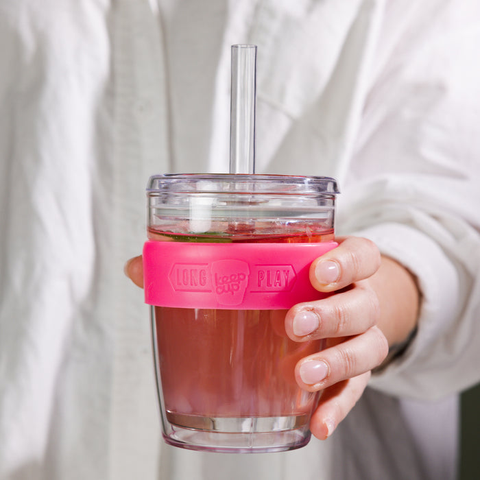 Hand holding a KeepCup Cold Cup Longplay Boba M 12oz in Flamingo, featuring a clear glass cup with pink silicone band, filled with iced tea and topped with a clear lid and reusable straw.