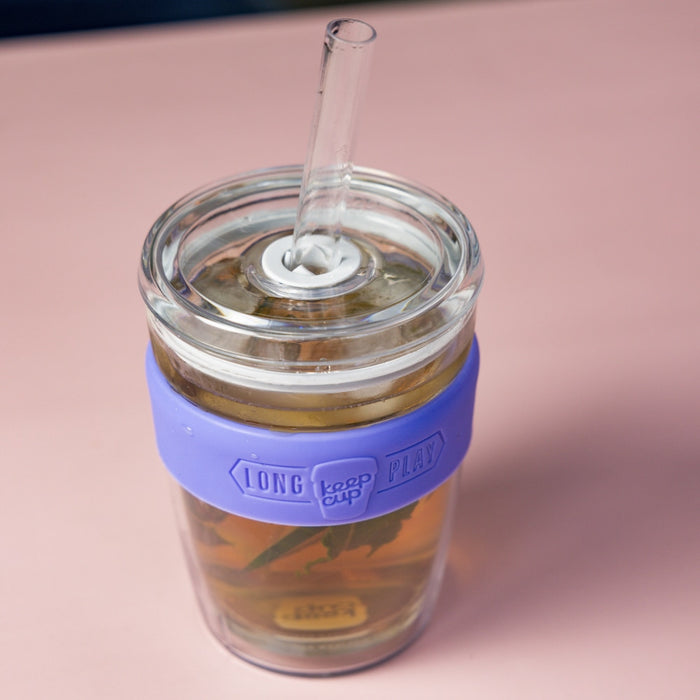 KeepCup Cold Cup Longplay Boba M 12oz in Indigo, shown from a top-down angle on a pink surface, featuring a clear glass straw inserted through the lid and filled with iced tea and visible tea leaves, with the lavender-coloured silicone band displaying the KeepCup Long Play logo.