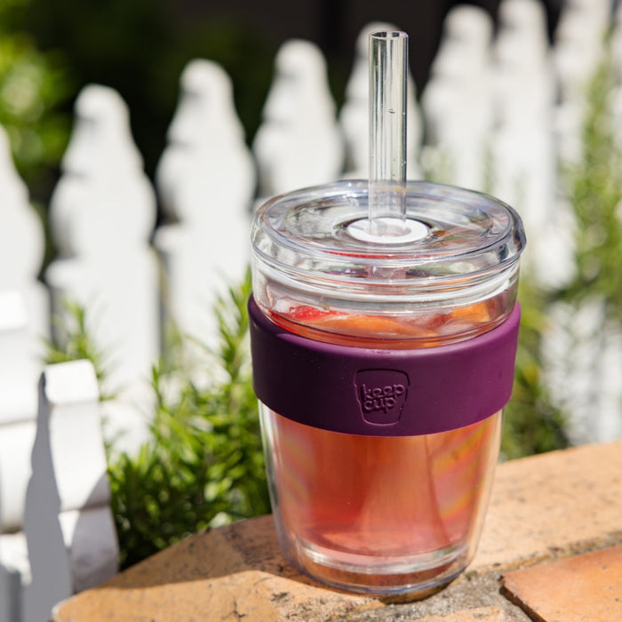 KeepCup Cold Cup Longplay Boba M 12oz in Merlot, featuring a glass cup with deep purple silicone band, clear lid and glass straw, filled with iced tea and fruit, sitting on a brick surface with a white picket fence and greenery in the background.