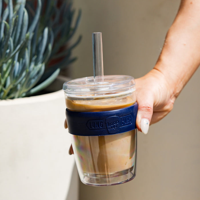 Hand holding a Cold Cup Longplay Boba M 12oz in Navy, filled with iced coffee and fitted with a clear straw, with a succulent plant in a white pot visible in the background.