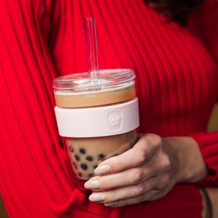 A woman in a red ribbed top holds a KeepCup Cold Cup Longplay Boba in Pashmak, featuring a clear glass body with pale pink silicone band, filled with bubble tea and tapioca pearls, with a reusable straw through the lid.