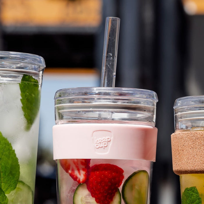 KeepCup Cold Cup Longplay Boba M 12oz in Pashmak, featuring a pink silicone band and clear glass body filled with strawberry and cucumber infused water, shown alongside other reusable cups on a café table with a glass straw inserted.