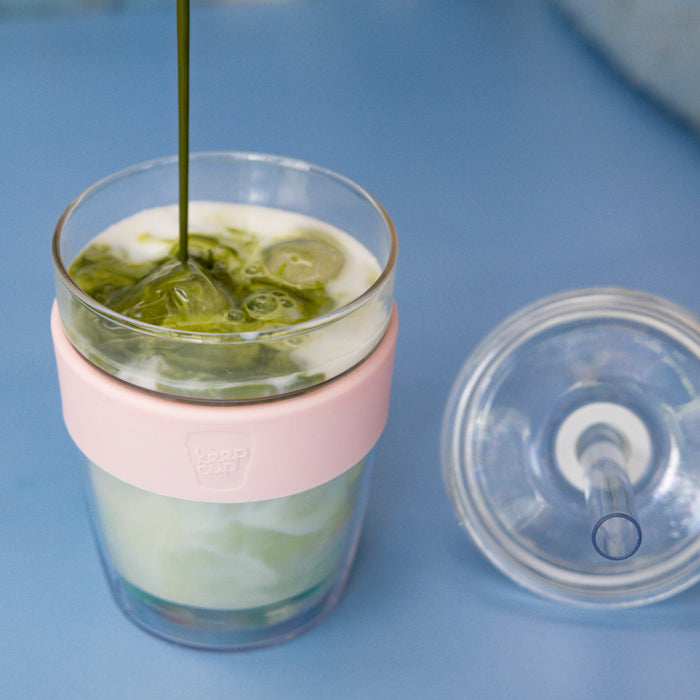 A KeepCup Cold Cup Longplay Boba in Pashmak sits on a blue surface with its lid removed, showing matcha being poured into milk and ice, with the pink silicone band and clear double-walled glass visible.