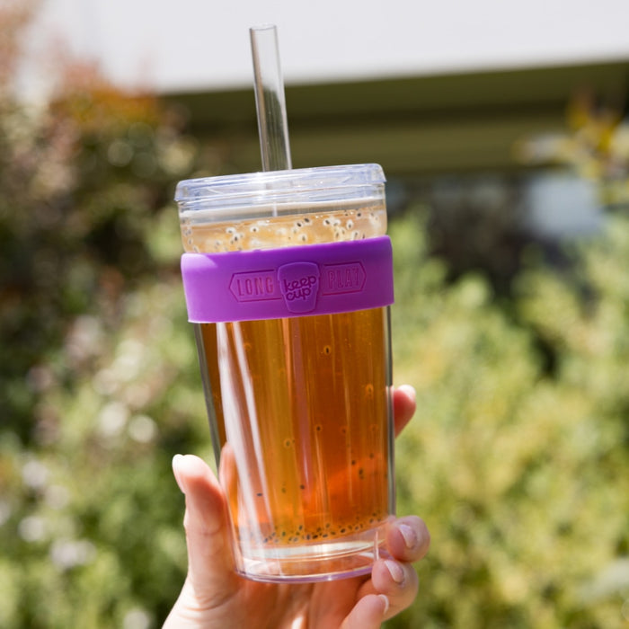 Hand holding a KeepCup Cold Cup Longplay Boba L 16oz in Bubblegum, filled with iced tea containing boba pearls, featuring a clear double-walled glass body, purple silicone band, clear lid and straw, photographed outdoors with greenery in the background.