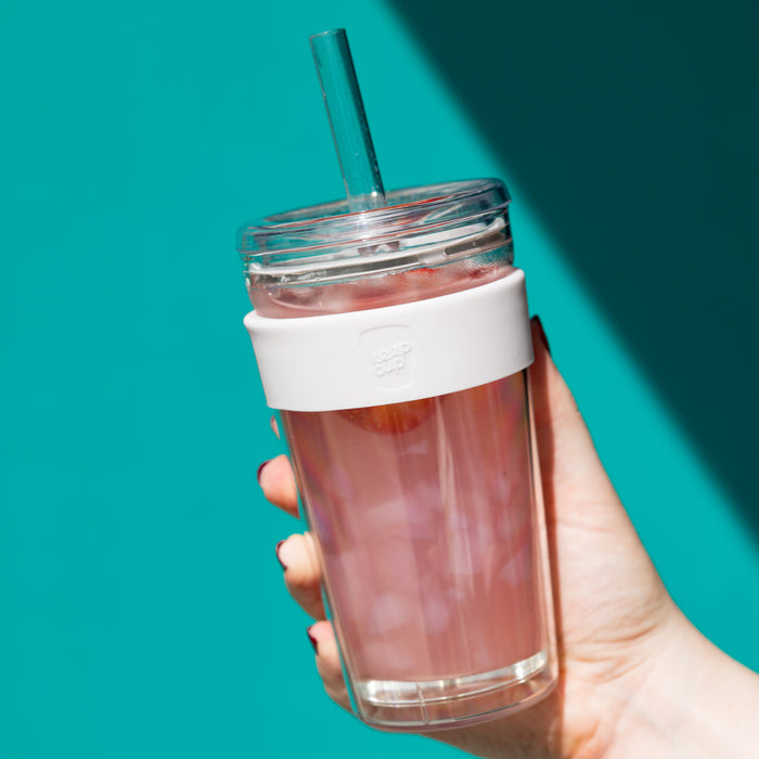 Hand holding a KeepCup Cold Cup Longplay Boba L 16oz in Chalk, filled with a pink iced beverage and ice cubes, featuring a clear lid with reusable straw and white silicone band, against a vibrant teal background.