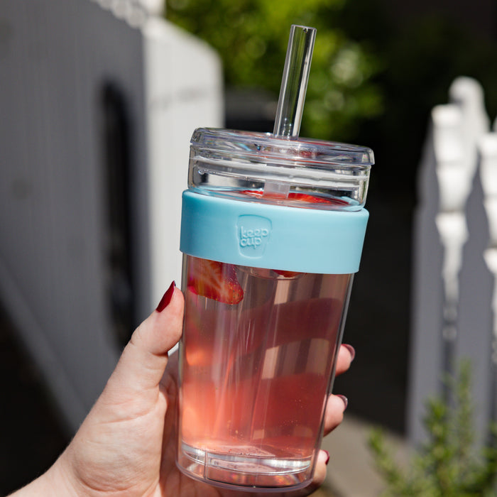 Hand holding a KeepCup Cold Cup Longplay Boba L 16oz in Cloud, filled with pink iced drink and strawberry slices, featuring a light blue silicone band and clear straw lid, photographed outdoors with a white fence in the background.