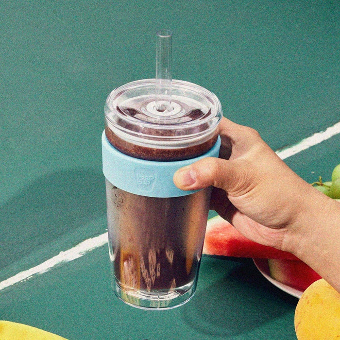 Hand holding a KeepCup Cold Cup Longplay Boba L 16oz in Cloud colour, filled with an iced chocolate drink and clear straw, set against a teal tennis court surface with watermelon slices and fruit visible in the background.