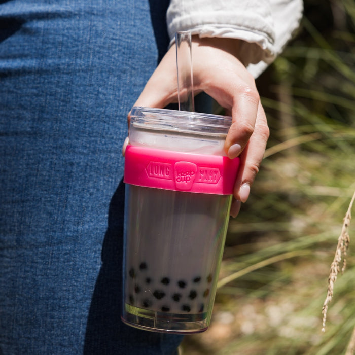 Hand holding a Cold Cup Longplay Boba L 16oz in Flamingo, filled with bubble tea and tapioca pearls, featuring a clear straw and pink silicone band, with Australian native grasses in the background.