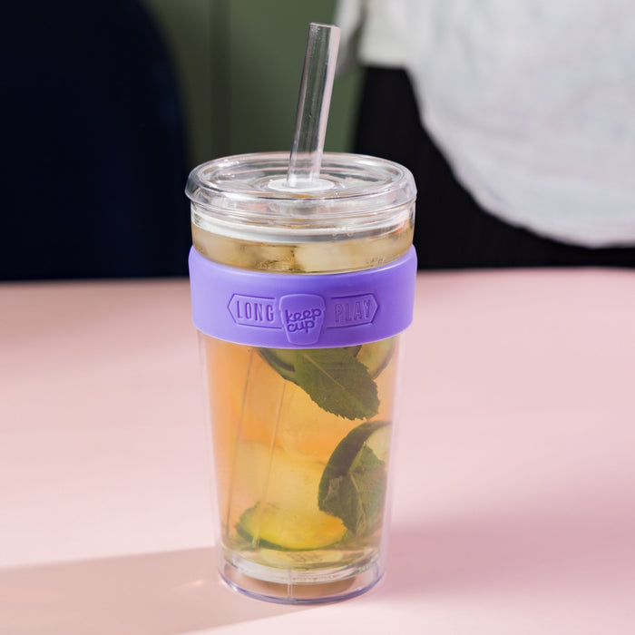 KeepCup Cold Cup Longplay Boba L 16oz in Indigo, featuring a clear double-walled cup with purple silicone band, filled with iced tea, mint leaves and citrus slices, shown with a glass straw on a pink surface.