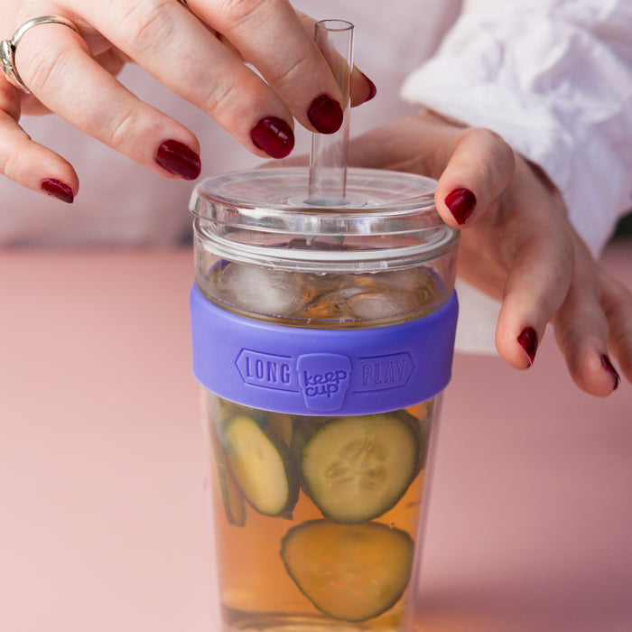A woman's hands with dark red nail polish hold a Cold Cup Longplay Boba L 16oz in Indigo, filled with iced tea and cucumber slices, with a clear straw inserted through the lid, set against a soft pink background.