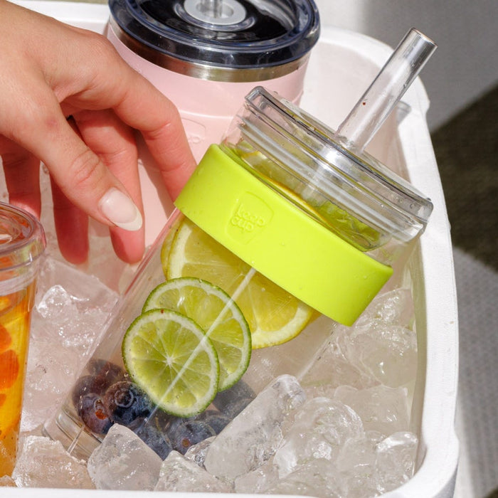 A hand reaches into an ice-filled cooler to grab a KeepCup Cold Cup Longplay Boba L 16oz in Matcha, featuring a lime green silicone band and clear body filled with citrus slices, blueberries and water, alongside other chilled beverages.