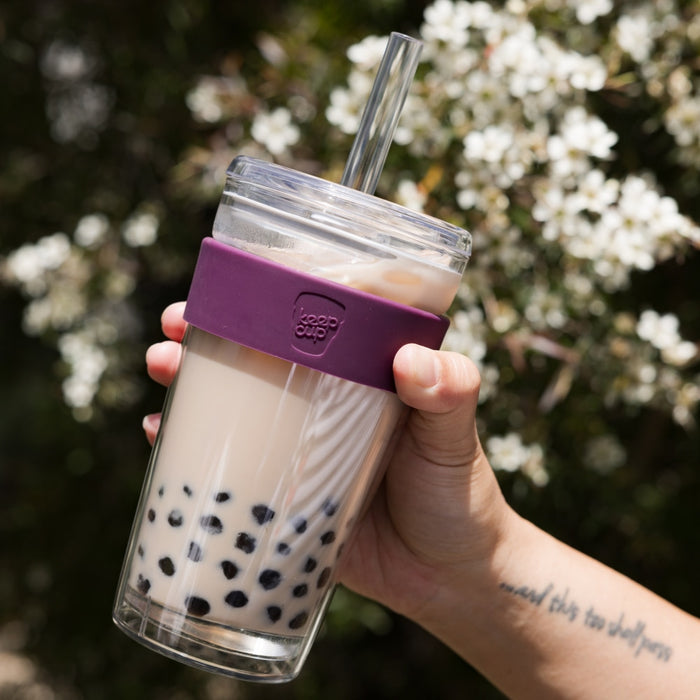 Hand holding a KeepCup Cold Cup Longplay Boba L 16oz in Merlot, filled with creamy bubble tea and dark tapioca pearls, featuring a purple silicone band and clear straw lid, photographed outdoors against white blossoms.