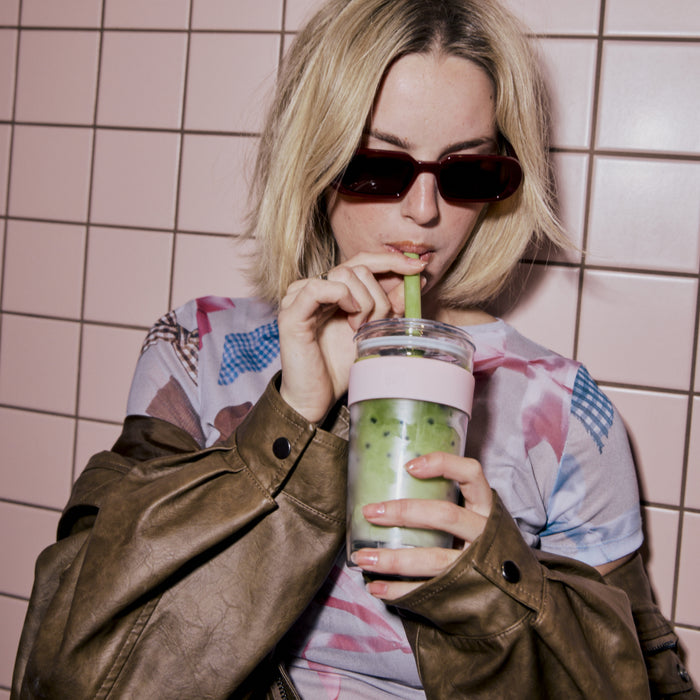 Woman with blonde hair and sunglasses drinking from a KeepCup Cold Cup Longplay Boba L 16oz in Pashmak, featuring a pale pink band and green iced beverage, against a pink tiled background.