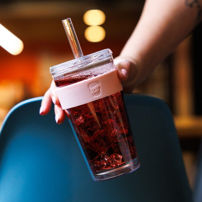 A hand with painted nails holds a KeepCup Cold Cup Longplay Boba L 16oz in Pashmak, filled with a dark red iced drink, against a blurred café background with warm bokeh lighting.