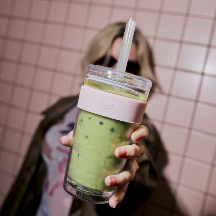 A person with long blonde hair holds a KeepCup Cold Cup Longplay Boba L 16oz in Pashmak towards the camera, filled with iced matcha and boba pearls, against a pink tiled wall backdrop.