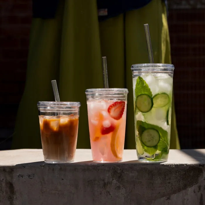 Three KeepCup Cold Cup Originals in different sizes arranged on a concrete ledge in sunlight, featuring an iced coffee with Coffee Straw in the M 12oz size, a strawberry lemonade, and a cucumber mint water, each with clear lids and reusable straws against a green fabric backdrop.