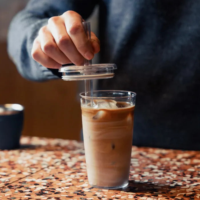A hand places a clear lid with straw onto a Cold Cup Original filled with iced coffee, set on a terrazzo café table with a ceramic cup visible in the background.