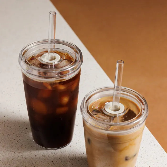 Two KeepCup Cold Cup Original L 16oz tumblers with clear lids and coffee straws, one filled with black iced coffee and the other with iced latte, positioned on a cream-coloured surface against a warm tan background.
