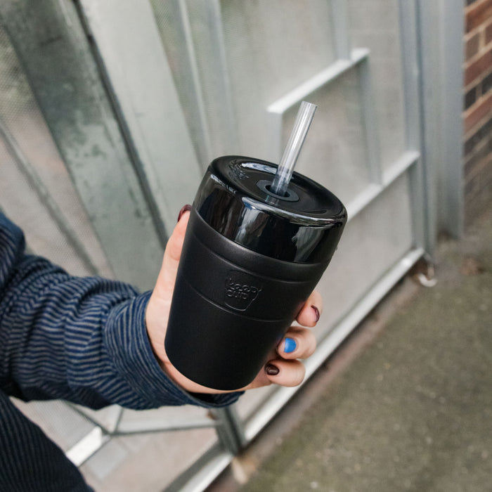 Hand with colourful painted nails holding a black KeepCup Commuter Cold Cup M 12oz with clear straw, photographed from above against an urban outdoor backdrop with metal stairs.