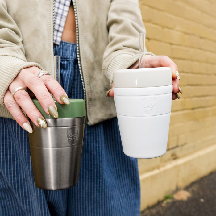 A person holding two KeepCup reusable cups outdoors, with the Commuter Cold Cup M 12oz in Bone displayed in their left hand featuring a white body with straw, while their right hand holds a stainless steel KeepCup with olive green lid.