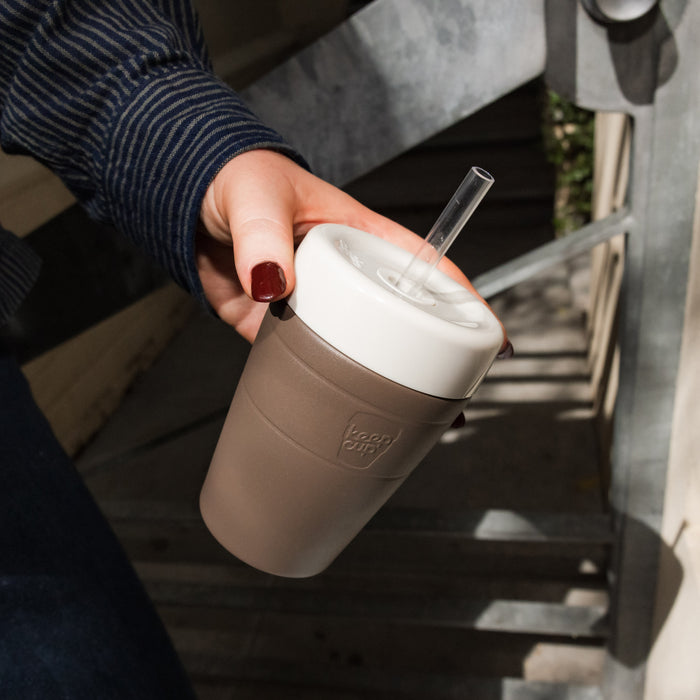A hand with dark red nail polish holds a KeepCup Commuter Cold Cup in Chocolate, featuring a white lid with clear straw, photographed from above on outdoor wooden stairs.