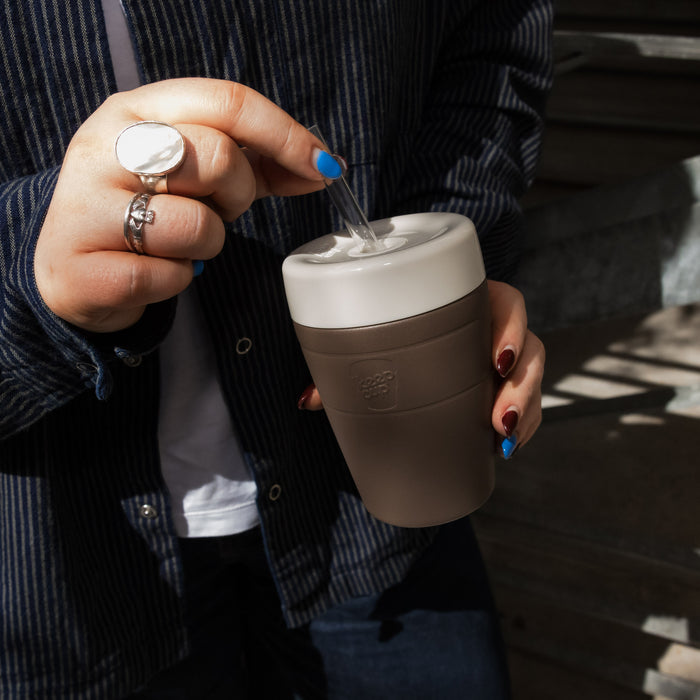 A person in a navy pinstripe jacket holds a KeepCup Commuter Cold Cup in Chocolate colour, inserting a clear straw through the white lid, showcasing the reusable cup's mid-size 12oz design with its taupe silicone band.