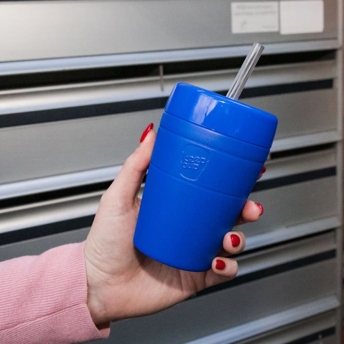 A hand with red nail polish holds the KeepCup Commuter Cold Cup in Electric blue with a clear straw, photographed against a grey louvred background.