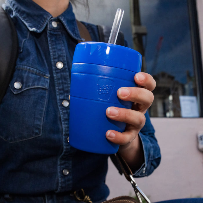 A hand holds the KeepCup Commuter Cold Cup M 12oz in Electric blue with a clear straw, worn casually with a denim jacket in an outdoor urban setting.