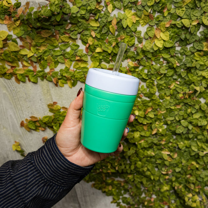 A hand with dark nail polish holds the KeepCup Commuter Cold Cup M 12oz in Emerald, featuring a bright green body, white lid, and clear straw, against a backdrop of green climbing leaves on a concrete wall.