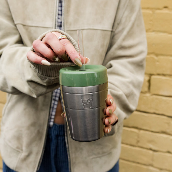 A person in a grey jacket holds a KeepCup Commuter Cold Cup in Pine, with a stainless steel body and sage green lid, inserting a clear straw into the silicone plug opening.