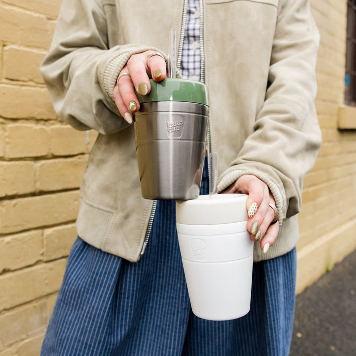 A person in a beige jacket holds two KeepCup reusable cups — a stainless steel Commuter Cold Cup in Pine with a clear straw in one hand, and a white cup in the other — against a yellow brick wall background.