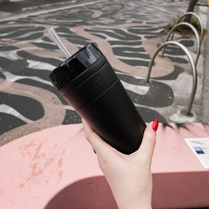 Hand with red nail polish holding a black KeepCup Commuter Cold Cup L 16oz with clear straw, against a backdrop of distinctive black and white wave-patterned pavement and pink outdoor seating.