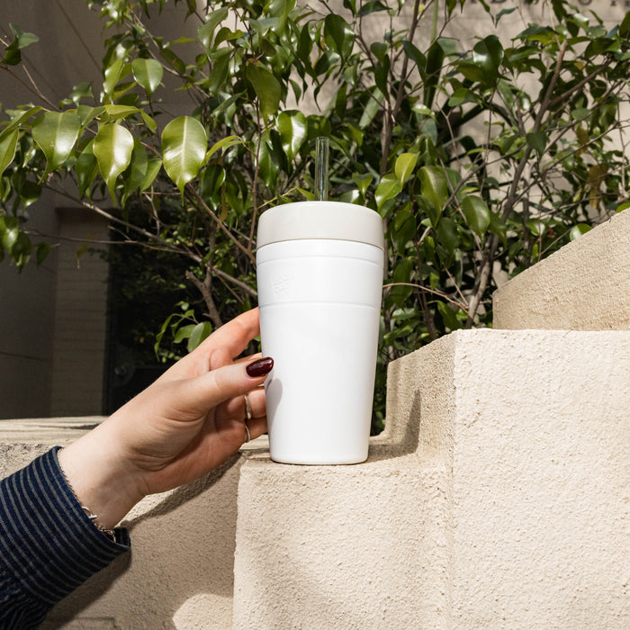 A hand with dark red nail polish reaches for a KeepCup Commuter Cold Cup in Bone, featuring a clear straw, positioned on a sunlit concrete ledge with green foliage in the background.
