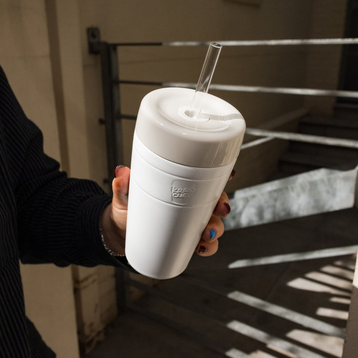 A hand with painted nails holds a KeepCup Commuter Cold Cup in Bone, featuring a clear straw and white lid, against an urban stairwell background with natural light casting shadows.