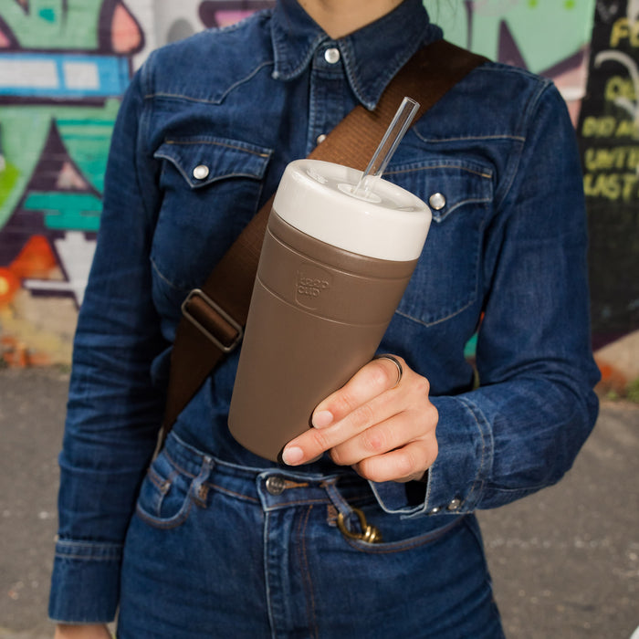 A person in a denim jacket holds a KeepCup Commuter Cold Cup L 16oz in Chocolate, featuring a taupe body with cream lid and clear straw, with colourful street art visible in the background.