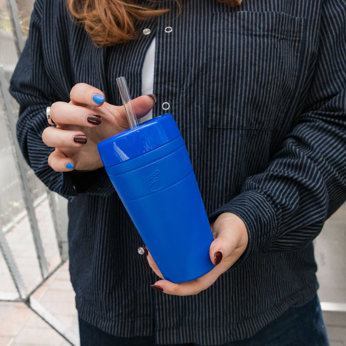 A person in a dark pinstriped jacket holds a KeepCup Commuter Cold Cup in Electric blue with a clear straw, showcasing the 16oz reusable cup's vibrant colour and comfortable grip design.