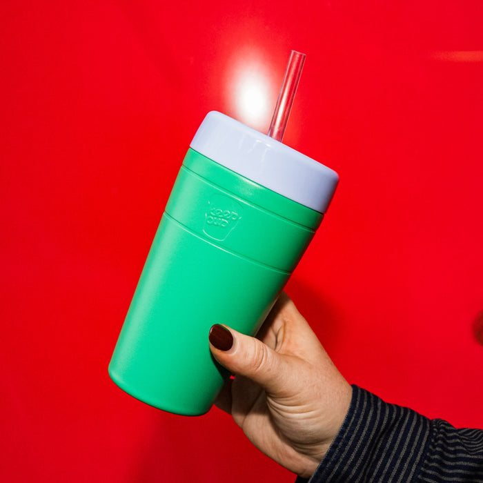 A hand with dark nail polish holds the KeepCup Commuter Cold Cup in Emerald, a vibrant green reusable cup with a grey lid and clear straw, photographed against a bold red background.