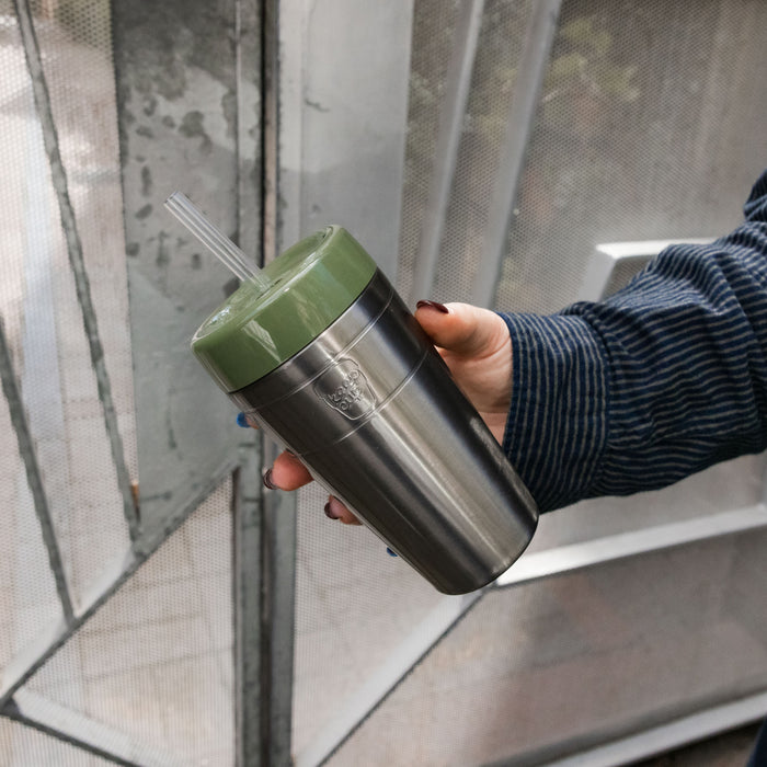 A hand with dark nail polish holds a KeepCup Commuter Cold Cup in Pine, featuring a stainless steel body and olive green lid with a clear straw, photographed at an angle near greenhouse panels.