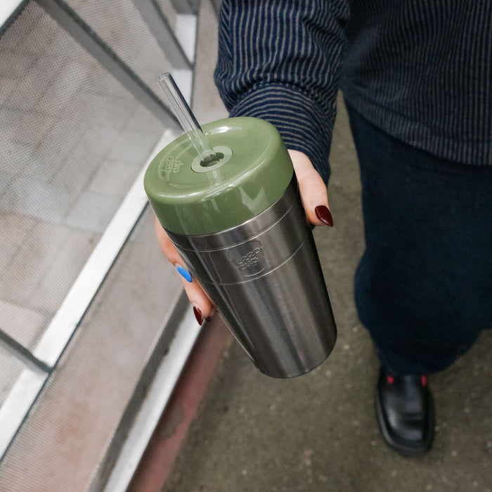A hand with colourful painted nails holds the KeepCup Commuter Cold Cup in Pine, featuring a stainless steel body and olive green lid with clear straw, photographed from above on a city street.