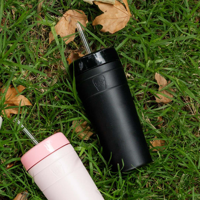 Two KeepCup Commuter Cold Cups lying on green grass with scattered autumn leaves — the XL 20oz Black cup with metal straw in the centre, alongside a smaller pink and white cup.