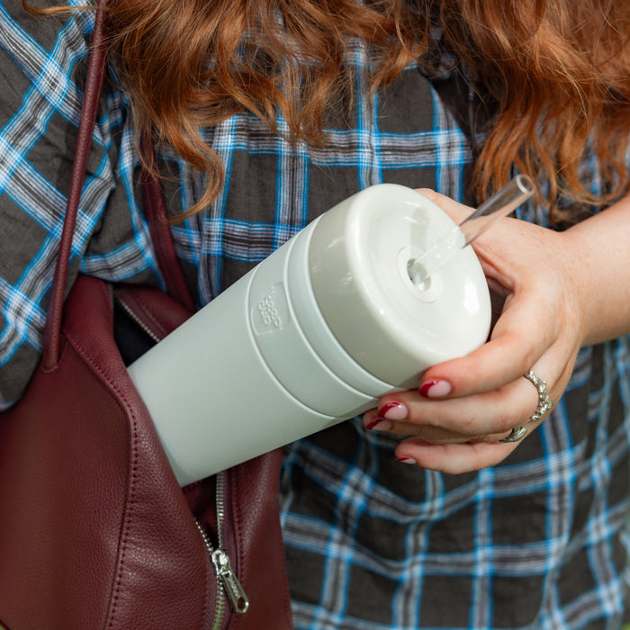 A woman with red curly hair wearing a grey and blue plaid shirt holds a KeepCup Commuter Cold Cup XL 20oz in Bone colour with both hands, a clear straw inserted in the lid, with a burgundy leather bag visible at her side.