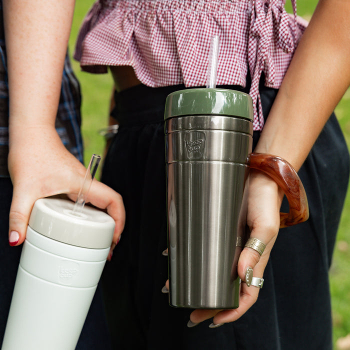 Two people outdoors holding KeepCup reusable cups, with one person displaying the Commuter Cold Cup XL 20oz in Bone featuring a straw, alongside a stainless steel KeepCup with olive green lid.
