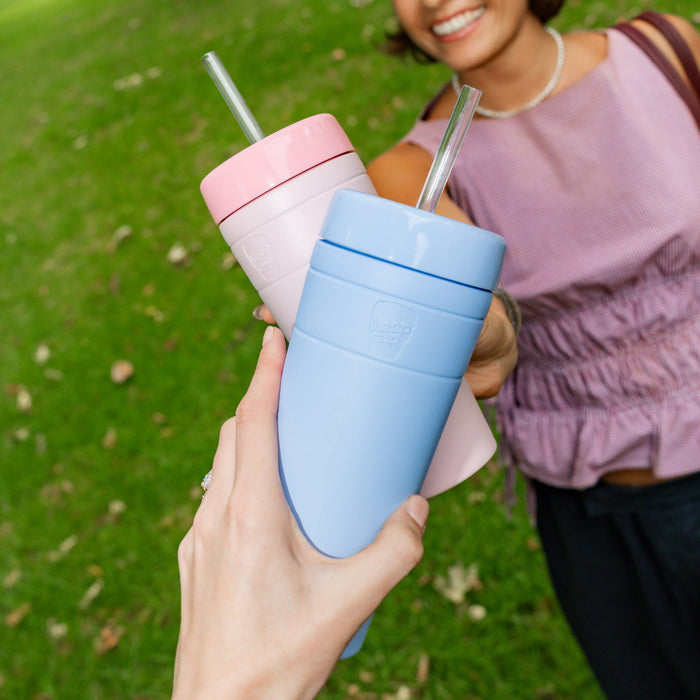 Two friends outdoors touching their KeepCup Commuter Cold Cups together in a cheerful toast, featuring the XL 20oz Orchid colour in soft pink and a light blue variant, both with straws, against a grassy park background.