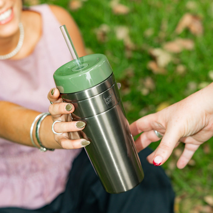 A woman in a pink top holds a KeepCup Commuter Cold Cup XL 20oz in Pine, featuring a stainless steel body and sage green lid with reusable straw, while seated outdoors on grass as another person reaches toward the cup.
