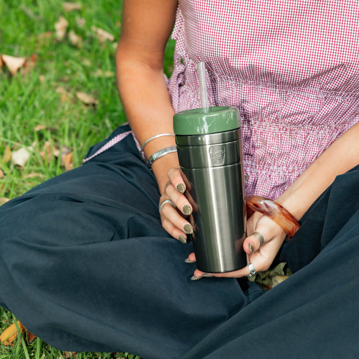 A person sitting cross-legged on grass holds a KeepCup Commuter Cold Cup XL 20oz in Pine, featuring a stainless steel body with olive green lid and clear straw, wearing a pink gingham top, dark trousers, and accessorised with silver bangles and rings.