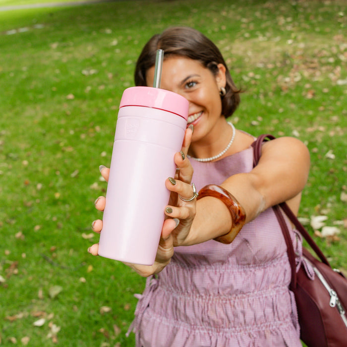 A smiling woman in a mauve dress extends her arm towards the camera, holding a KeepCup Commuter Cold Cup XL 20oz in Pashmak Pink with a metal straw, photographed outdoors in a park setting with green grass in the background.