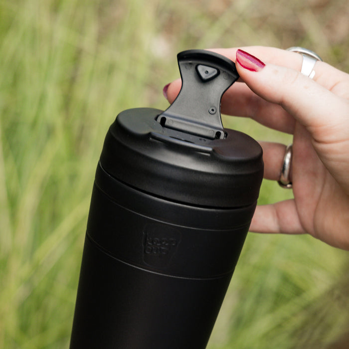 A hand with red nail polish opens the flip-top lid of a black KeepCup Commuter M 12oz reusable cup, with blurred green grass in the background.