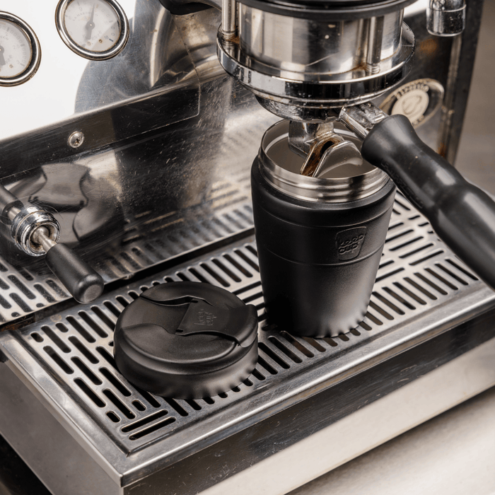 KeepCup Commuter M 12oz in Black positioned under an espresso machine group head with lid removed, ready for coffee to be poured directly into the reusable cup, with the black lid resting on the stainless steel drip tray beside it.